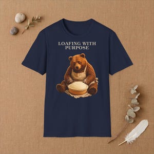 Könnte beinhalten: Ein marineblaues T-Shirt mit einer Grafik eines braunen Bären, der eine beige Schürze trägt und Teig in einem Gärkörbchen knetet. Der Text "LOAFING WITH PURPOSE" ist in weißen Großbuchstaben über der Bärenillustration auf der Vorderseite gedruckt.