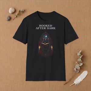 Può includere: Una t-shirt nera a maniche corte con la scritta bianca BOOKED AFTER DARK sopra l'immagine di un pipistrello che legge un libro all'interno di un arco gotico, affiancato da due candele accese e rose rosse sotto una luna crescente nel cielo notturno.