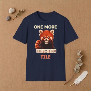 May include: A navy blue t-shirt featuring a graphic of a red panda holding a mahjong tile. Above the panda, the text reads ONE MORE, and below a row of mahjong tiles, the text reads TILE. The design uses cream, red, and orange colours.