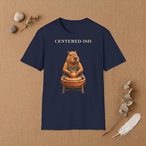 Könnte beinhalten: Ein dunkelblaues kurzärmeliges T-Shirt mit einer Grafik eines braunen Wasserschweins an einer Töpferscheibe. Über dem Wasserschwein steht der Text CENTERED-ISH in weißen Großbuchstaben. Das Shirt hat einen Rundhalsausschnitt und einen geraden Saum.