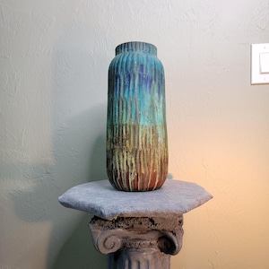 May include: A tall, cylindrical ceramic vase featuring a vertical ribbed texture. The glaze transitions from a deep teal and blue at the top to a muted olive green and earthy brown at the base, creating a gradient effect across the surface.