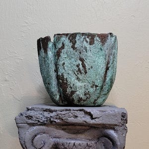 May include: A textured, cup-shaped vessel with a weathered, oxidized teal and brown patina finish. The vessel features a rugged, uneven surface and sits atop a gray, stone-like pedestal base with a classic architectural scroll design.