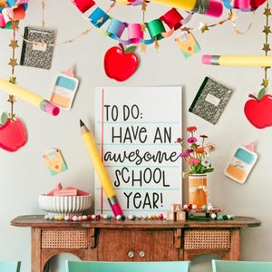 以下が含まれることがあります： 青い水平線と黒い文字でTO DO: HAVE AN AWESOME SCHOOL YEAR!と書かれた白い長方形の看板。周囲には紙のチェーン、大きな黄色い鉛筆の切り抜き、赤いリンゴの形、糊のボトル、ノートが飾られている。