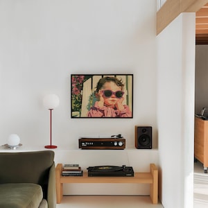 May include: A rectangular framed wall art print featuring a stylised illustration of a child with curly hair wearing bright pink sunglasses and a pink collared shirt, positioned against a backdrop of white fencing and green foliage.