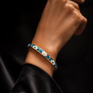 Peut inclure: Un bracelet jonc articulé doré avec un fond en émail bleu sarcelle vif, orné de motifs floraux blancs et de détails en forme de feuilles dorées. Le bracelet présente une finition lisse et polie ainsi qu'un profil circulaire fin.
