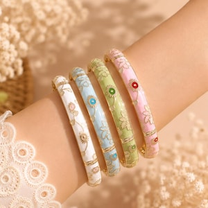 Peut inclure: Quatre bracelets joncs articulés en émail blanc, bleu clair, vert clair et rose clair. Chaque bracelet présente un motif floral doré avec de petits accents circulaires colorés au centre des fleurs, disposés en rangée sur un bras.