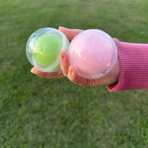 May include: Two spherical, translucent plastic containers, each holding a smooth, solid-colored substance. One container holds a bright lime green sphere, while the other contains a soft pastel pink sphere, both featuring a clear, hinged shell.