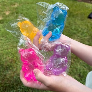 Könnte beinhalten: Vier einzelne bärenförmige Objekte aus einem durchscheinenden, geleeartigen Material, die jeweils in eine durchsichtige Plastikfolie eingewickelt sind. Die Bären sind leuchtend gelb, blau, pink und violett und haben glatte Konturen.