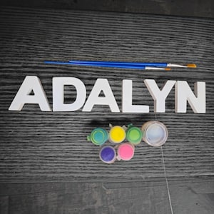 May include: White wooden letters spelling the name ADALYN, accompanied by two blue-handled paintbrushes and a set of six small circular paint pots containing yellow, green, blue, purple, pink, and white paint colors for craft projects.
