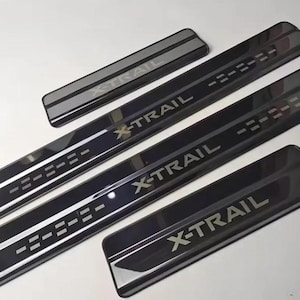 May include: A set of four black door sill protectors for a vehicle, featuring a glossy finish and the text X-TRAIL in silver lettering. Two long strips include a dashed line pattern, while two shorter rectangular pieces feature the same logo.