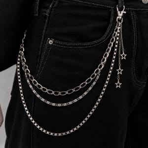 Puede incluir: Una cadena de billetera de varias capas en tono plateado sujeta a pantalones vaqueros negros. El accesorio presenta tres estilos de cadena distintos, incluidos eslabones ovalados y una cadena cuadrada, con tres dijes de estrella.