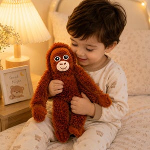 Puede incluir: Un peluche de orangután con pelaje largo de color naranja oscuro, cara clara y ojos grandes y oscuros. El animal de peluche presenta brazos y piernas largos, una textura suave y una expresión amigable, diseñado para el confort y el juego.