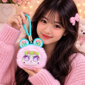 May include: A round, fluffy pink plush pouch featuring a white face with large, detailed purple eyes, a small nose, and a floral crown. The pouch has two teal ears decorated with flowers and a matching teal braided wrist strap attached to the top.