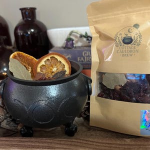 May include: A black cast iron cauldron with a triple moon symbol, filled with dried orange slices and bay leaves, next to a tan pouch labeled Cottage Cauldron Brew containing dried botanicals and a holographic sticker reading The Apothecary Aching.