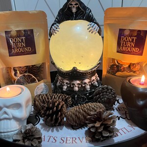 May include: A black tray holds two beige pouches labeled DON'T TURN AROUND, a glowing crystal ball held by a grim reaper figure with a skull base, a white skull candle holder, a black skull candle holder, and several brown pine cones.