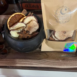 Op de afbeelding: Een zwarte ketel gevuld met gedroogde sinaasappelschijfjes, appelschijfjes en steranijs, naast een bruin papieren zakje met het label Midnight Orchard met gedroogd fruit en kaneelstokjes, voorzien van een holografische sticker rechtsonder.
