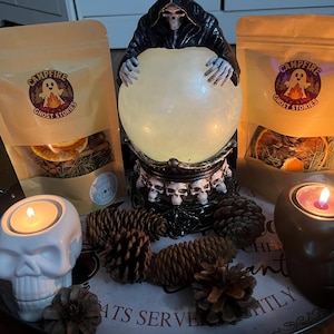 Peut inclure: Deux sachets beiges étiquetés Campfire Ghost Stories contenant des fruits séchés et des aiguilles de pin, une boule de cristal lumineuse tenue par une faucheuse, un bougeoir crâne blanc, un bougeoir crâne noir et des pommes de pin sur un plateau.