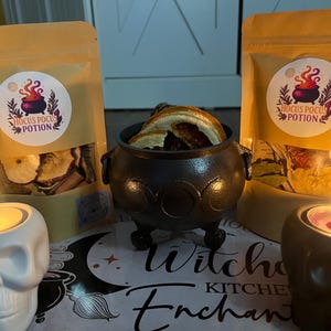 May include: Two tan pouches labelled Hocus Pocus Potion with dried fruit, a black three-legged cauldron filled with dried orange slices, and two skull-shaped candle holders with lit tea lights, all resting on a mat with the text Witch's Kitchen Enchantment.