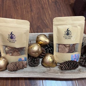May include: Two tan stand-up pouches labelled Gilded Pear with a witch hat and pear graphic, arranged in a wooden bowl with several metallic gold-painted pears and natural brown pinecones. The pouches contain dried food items visible through a clear window.