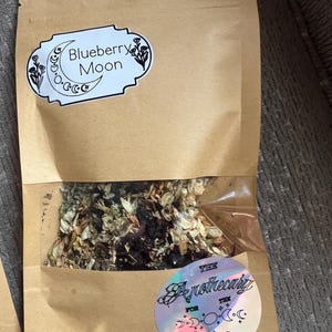 Puede incluir: Una bolsa de papel marrón con una ventana transparente que contiene una mezcla de hierbas secas. La etiqueta superior dice Blueberry Moon con un gráfico de luna creciente. La etiqueta holográfica circular inferior dice The Apothecary for the Aching.