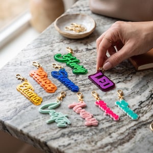 May include: A collection of colorful acrylic name keychains featuring various names including Eliana, Luca, Henry, Leo, Carter, and Avery. Each keychain has a gold-toned metal lobster clasp and is crafted from layered, translucent or solid acrylic.