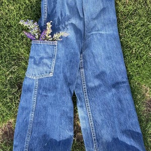 May include: A pair of wide-leg blue denim trousers featuring a single patch pocket on the front left side. The pocket contains a small bouquet of purple lavender sprigs. The denim fabric has a medium wash with visible stitching along the seams.