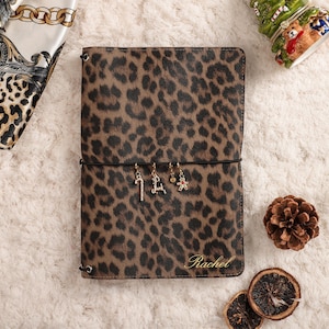 May include: A rectangular notebook with a brown and black leopard print cover. The name Rachel is embossed in gold in the bottom right corner. A black elastic band wraps around the center, holding four small metal charms: a candy cane, a letter L, a small bead, and a gingerbread man.