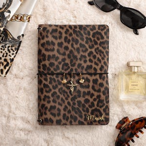 May include: A rectangular notebook with a brown and black leopard print cover featuring the name Mariah in gold script. A black elastic band holds three gold charms: a shell, a cross, and a heart. Nearby are black sunglasses, a perfume bottle, and a hair clip.