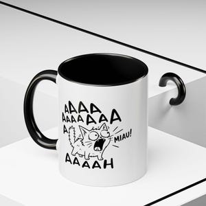 May include: A white ceramic mug with a black interior and a black handle. The exterior features a black line drawing of a wide-eyed, screaming cat with the text AAAAA AAAAAAA AAAAAH and MIAU printed in a bold, hand-drawn style font.