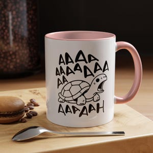 May include: A white ceramic mug with a soft pink interior and handle. The exterior features a black line drawing of a turtle with its mouth wide open, surrounded by the repeated text "AAAA AAAA DAA AA AADAAH" in a hand-drawn style font.