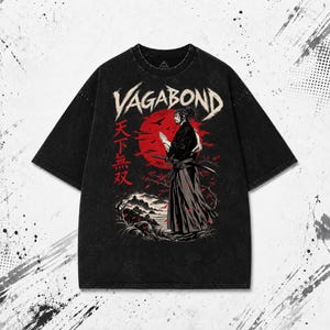 Peut inclure: Un t-shirt noir à manches courtes avec une impression graphique représentant un samouraï avec un sabre devant un grand soleil rouge. Le texte VAGABOND est imprimé en blanc au-dessus, avec des caractères japonais rouges sur la gauche.