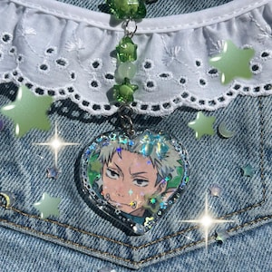 May include: A heart-shaped acrylic keychain featuring an anime character illustration with spiky hair. The charm has a holographic border and is attached to a silver metal ring by a chain of green star-shaped beads and a single round white bead.