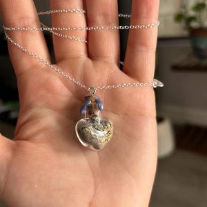 May include: A heart-shaped glass vial pendant on a thin silver-tone chain. The vial is filled with dried botanical material and sealed with a cork stopper topped with a small amount of light purple wax. The chain is delicate and metallic.