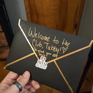 May include: A black envelope with gold trim featuring handwritten gold text that reads Welcome to the Club, Tabby! So glad you are here! and a small white sticker with a red heart and the text Rely ON JESUS centered on the envelope flap.