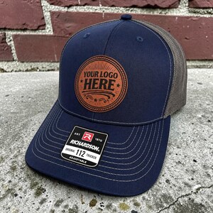 May include: A navy blue trucker hat with a brown circular leather patch featuring the text YOUR LOGO HERE. The hat has a mesh back, a curved brim with white stitching, and a black Richardson 112 Original adjustable sticker on the front brim.