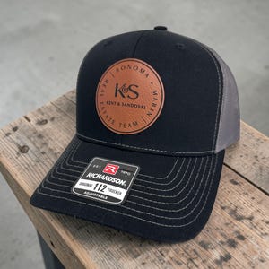May include: Black trucker hat with a gray mesh back and a curved brim. A brown leather patch features the text "SONOMA REAL ESTATE TEAM" and the initials "K&S". The hat has a "RICHARDSON" tag.
