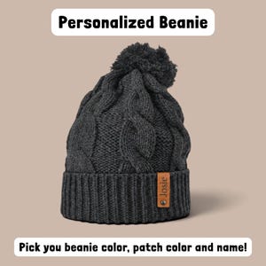 May include: A dark gray cable-knit beanie with a matching pom-pom on top. The beanie features a ribbed cuff with a tan rectangular leather patch attached by a metal rivet. The patch is embossed with the name Josie. Text reads Personalized Beanie.