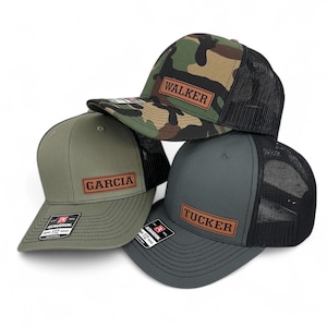 May include: Three trucker hats with leather patches. The hats are in camouflage, green, and gray. The patches have the names "Walker", "Garcia", and "Tucker" on them.