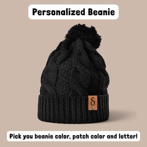May include: A black cable-knit beanie with a matching black pom-pom on top. The beanie features a ribbed cuff with a tan rectangular leather patch displaying the letter S and a small metal rivet. Text reads: Personalized Beanie. Pick you beanie color, patch color and letter!