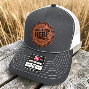 May include: A charcoal gray Richardson 112 trucker hat with a white mesh back. The front features a brown leather patch with the text "YOUR LOGO HERE" in a circular design. The hat has a curved brim with white stitching.