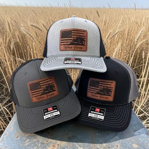 May include: Three trucker hats in gray, charcoal, and black, stacked on top of each other. Each hat features a brown leather patch with an American flag design and a tractor. The text "Weiss Farms" is below the flag.