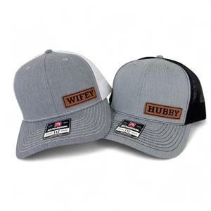 May include: Two gray trucker hats with leather patches. One hat has a white front panel and the word "Wifey" on the patch. The other hat has a black mesh back and the word "Hubby" on its patch. Both hats have the Richardson 112 tag.