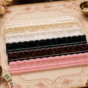 May include: A set of six rectangular mahjong tile racks made of acrylic in various colours and patterns, including clear, pearlescent white, marbled green, tortoiseshell brown, and marbled pink, each featuring a scalloped top edge design.