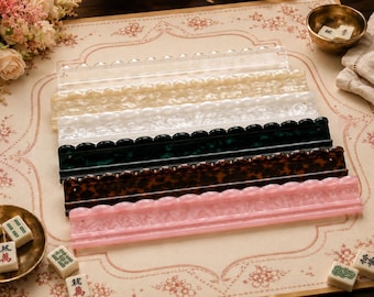 Puede incluir: Un conjunto de seis soportes rectangulares para fichas de mahjong hechos de acrílico en varios colores y patrones, incluyendo transparente, blanco perlado, verde jaspeado, marrón carey y rosa jaspeado, con un borde superior festoneado.