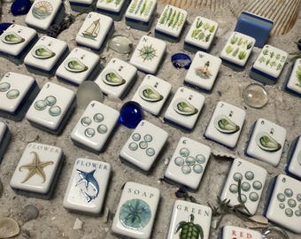 May include: A collection of rectangular white mahjong tiles featuring various coastal and nautical illustrations, including oysters, sailboats, lighthouses, starfish, sharks, sea turtles, lobsters, and botanical ferns, arranged on a sandy surface.