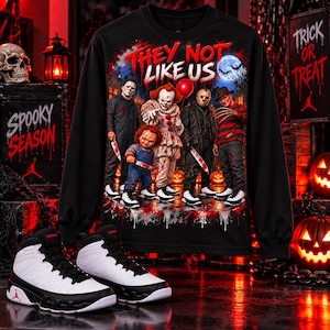 May include: A black long-sleeve shirt featuring a graphic print of horror movie characters and the text "THEY NOT LIKE US" above them. The design includes Michael Myers, Pennywise, Chucky, Jason Voorhees, and Freddy Krueger in a spooky setting.