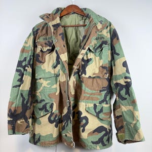 May include: A camouflage field jacket featuring a woodland pattern in shades of green, brown, and black. The jacket includes a front zipper, snap button closures, four large flap pockets, and the text WALKER and ARMY printed on the chest area.
