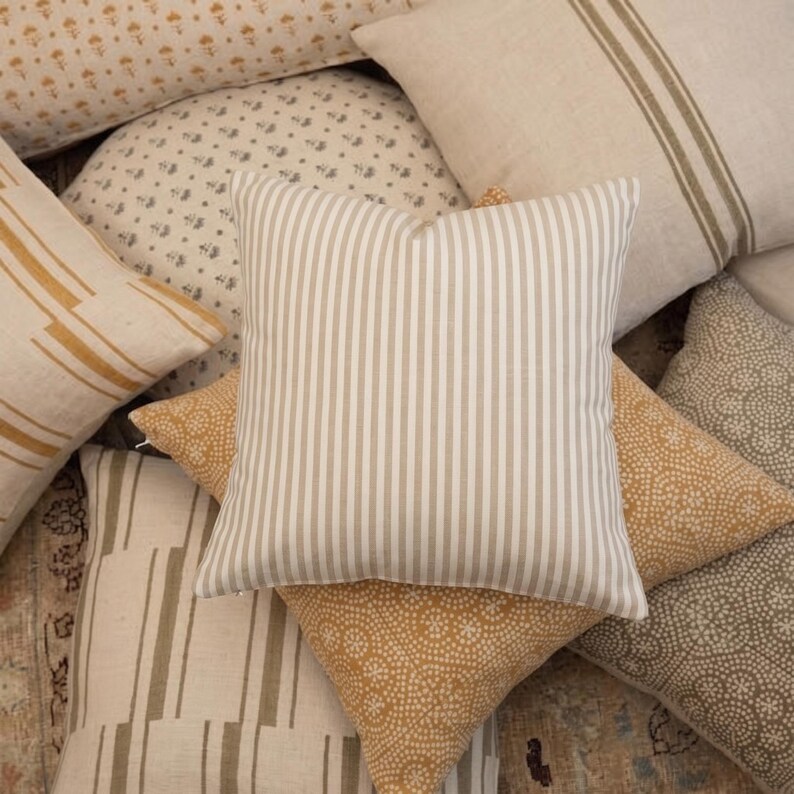 A close-up pile of various throw pillows featuring neutral patterns, dots, and stripes in beige, cream, and mustard tones.