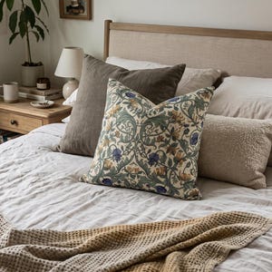A bed features light neutral linens, a textured tan throw blanket, and a collection of accent pillows with a patterned botanical pillow in front.