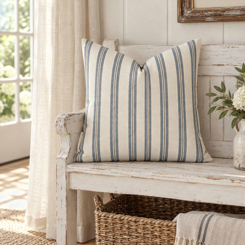 Slate blue and ivory ticking stripe throw pillow on a white wooden entryway bench with a woven basket underneath.